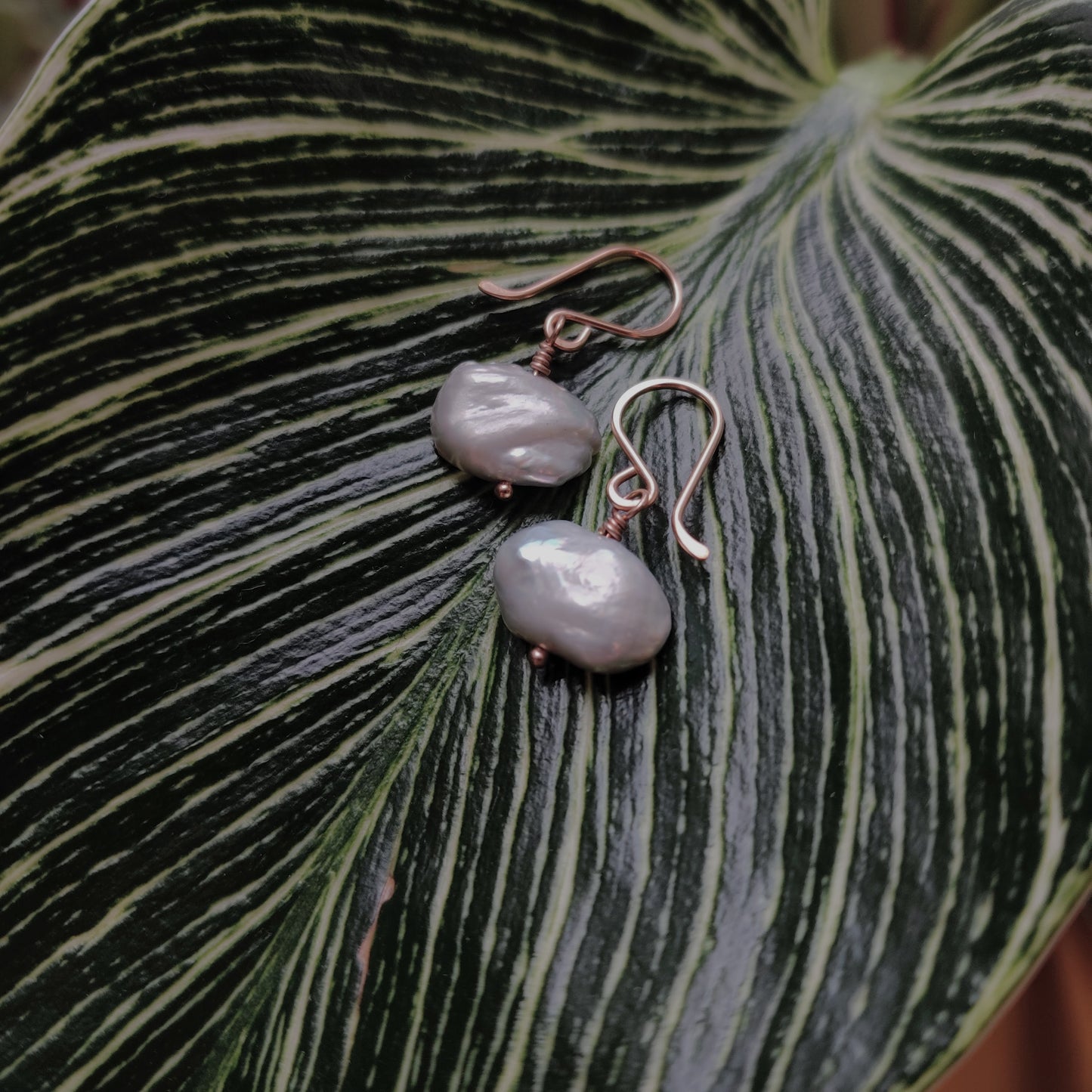 Flatlay image of Berlin jewelry designer Zurina Ketola's Organic Gray Pearl Drop Earrings in 14K rose gold fill laying on a large vareigated leaf.