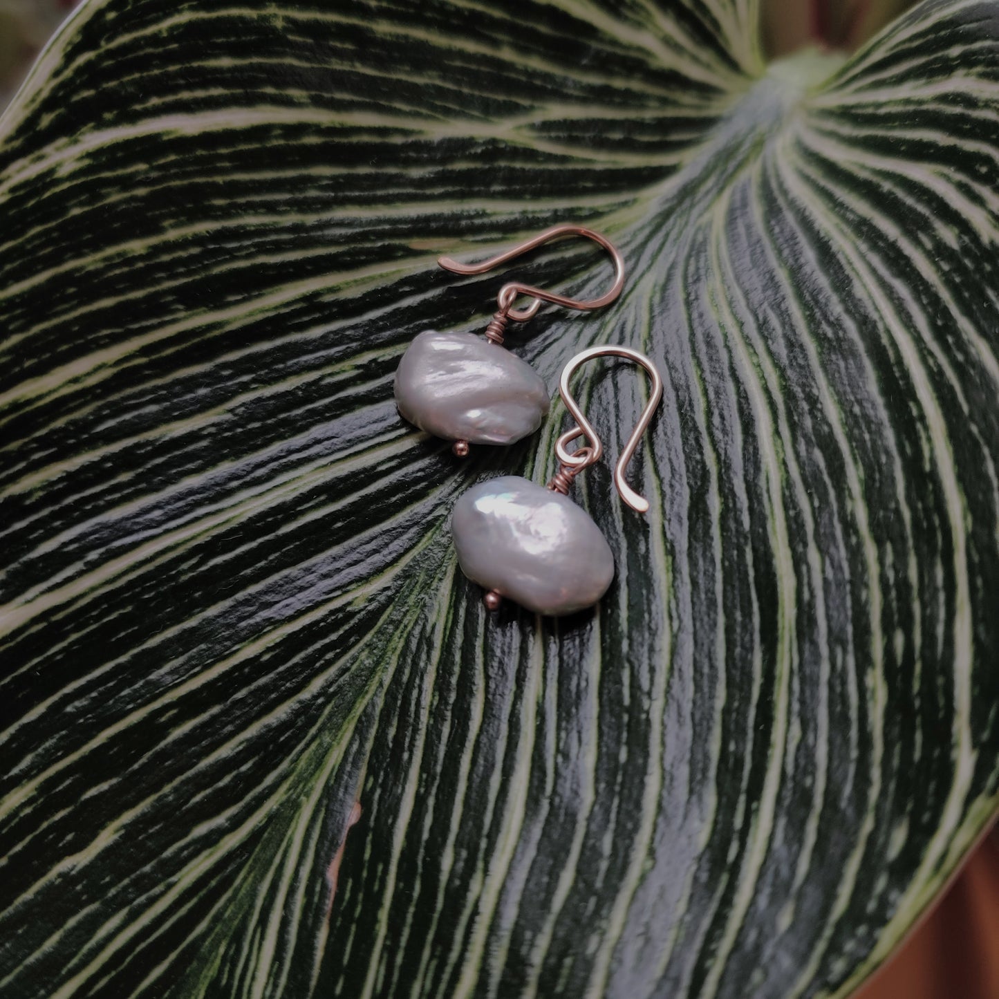 Flatlay image of Berlin jewelry designer Zurina Ketola's Organic Gray Pearl Drop Earrings in 14K rose gold fill laying on a large vareigated leaf. 